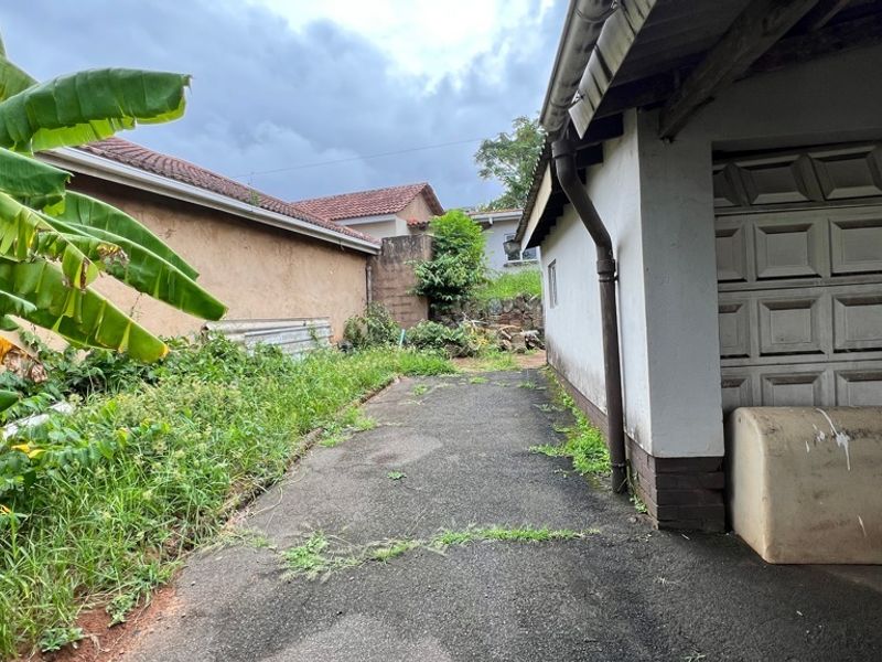 Rear exterior area featuring an asphalt driveway, garage access, and boundary wall, currently requiring significant landscaping and maintenance.
