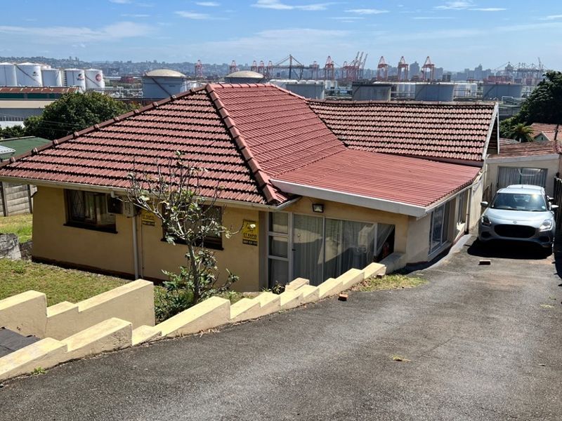 Front exterior view of a single-story house with a tiled roof, asphalt driveway, and retaining wall, overlooking an industrial harbor area.