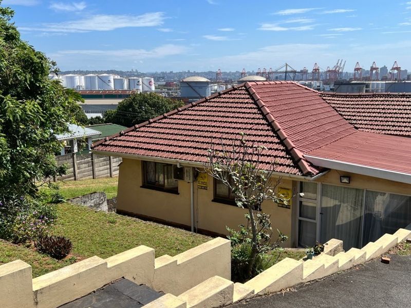 Rear view of the property featuring a tiled roof, sliding glass doors, and a tiered retaining wall overlooking an industrial harbor area.