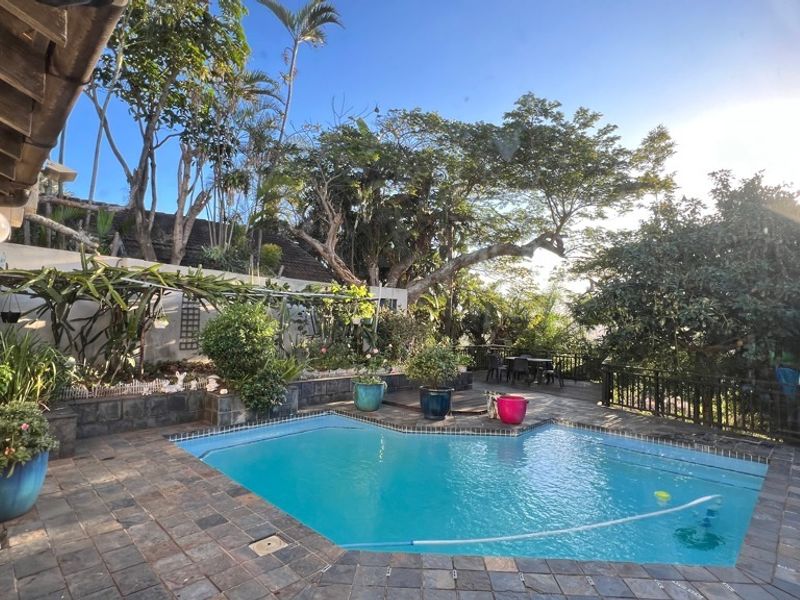 A private outdoor pool area featuring stone paving, a retaining wall, and a covered patio space surrounded by established garden landscaping.