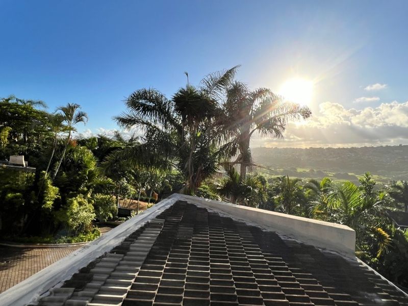 Elevated view overlooking a lush garden and surrounding landscape, featuring a tiled roof and parapet wall in the foreground.