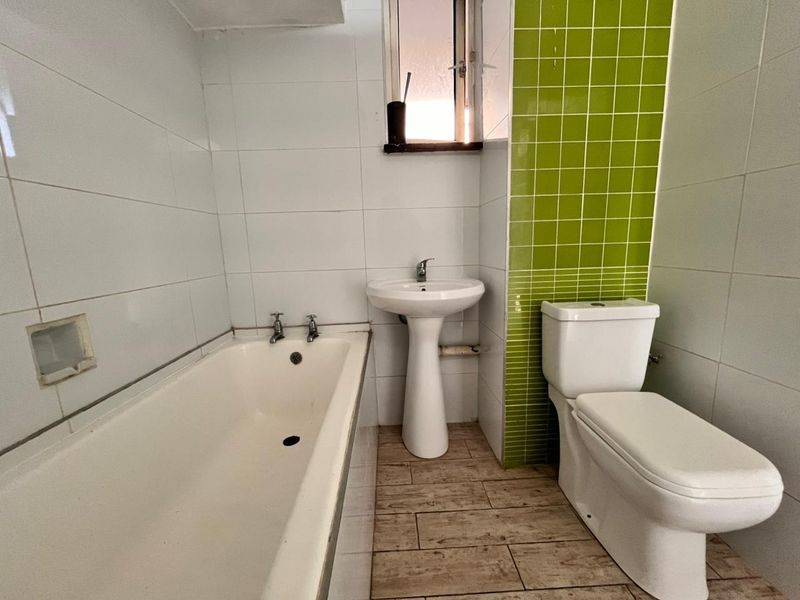 A functional bathroom featuring a bathtub, pedestal basin, and toilet, finished with white and green accent wall tiles and wood-look floor tiles.