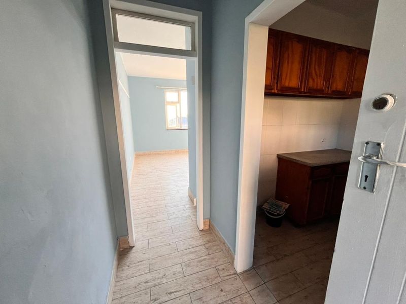A view from a hallway looking into a bedroom and a kitchen area featuring wooden cabinetry and tiled flooring throughout.