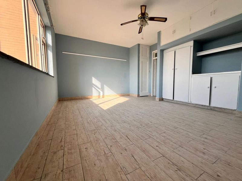 A bright, empty bedroom featuring wood-look tiled flooring, built-in storage cupboards, and a ceiling fan, offering a clean and neutral space.