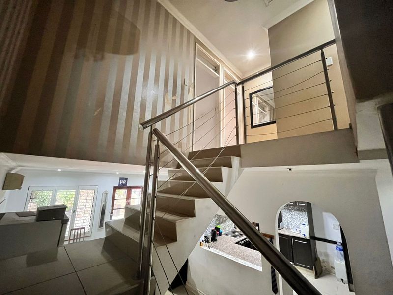 A central hallway featuring a modern stainless steel staircase railing, tiled flooring, and an archway leading to the kitchen area.