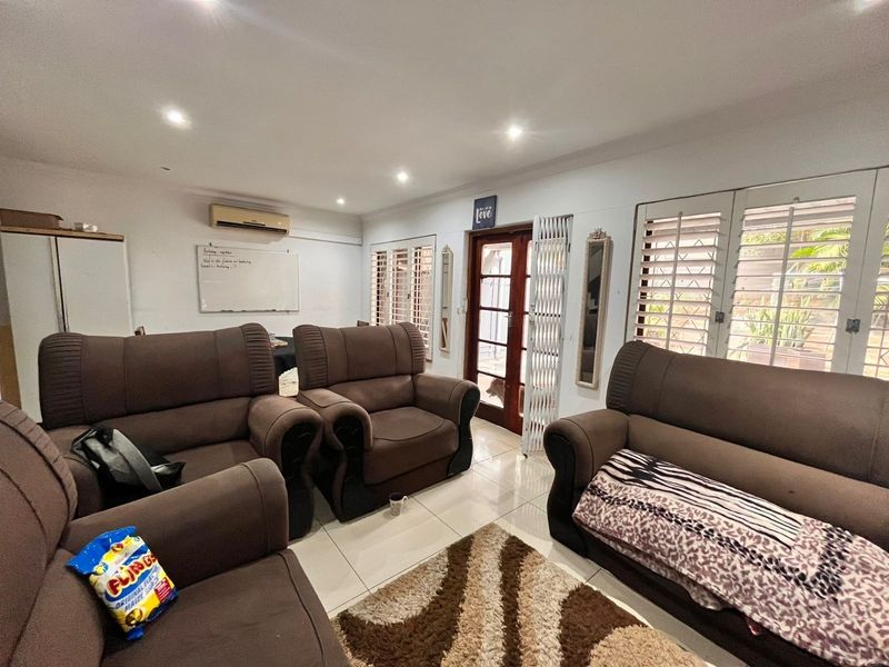 A spacious living room featuring tiled flooring, recessed downlighting, a wall-mounted air conditioner, and access to the exterior via a security-gated timber door.