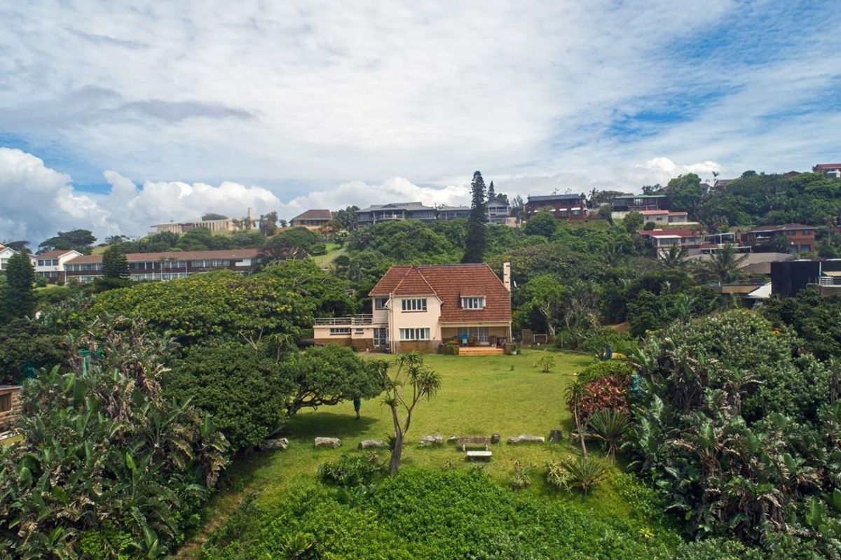 Aerial view of a detached house with a tiled roof, set within a large, lush garden surrounded by mature trees and coastal vegetation.