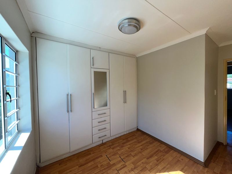 A bright, empty bedroom featuring built-in white cupboards with integrated drawers and mirror, complemented by warm-toned laminate flooring and natural light.