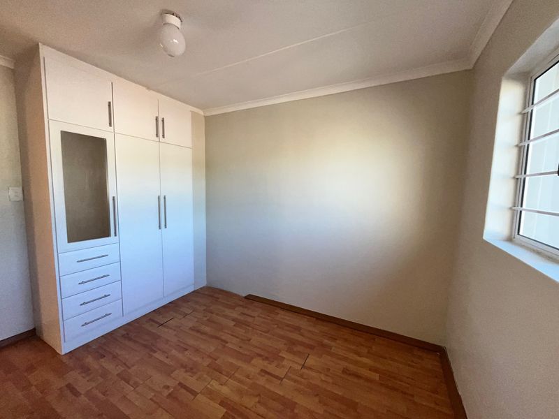 A bright, empty bedroom featuring built-in white cupboards, laminate flooring, and a window fitted with security bars.