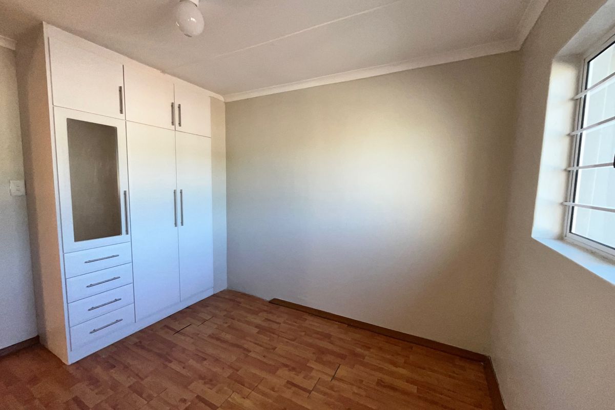 A bright, empty bedroom featuring built-in white cupboards, laminate flooring, and a window fitted with security bars.
