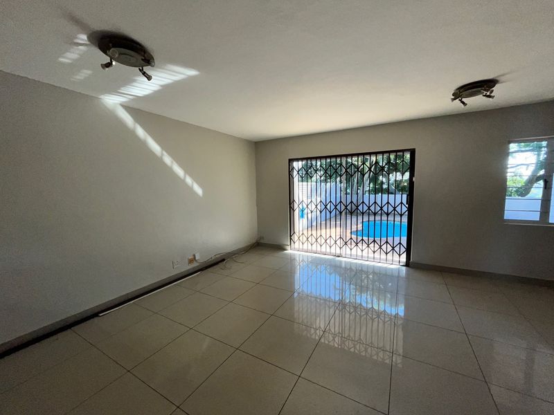 A bright, empty living room featuring large format floor tiles, ceiling lighting, and a sliding glass door with a security gate leading to the pool area.