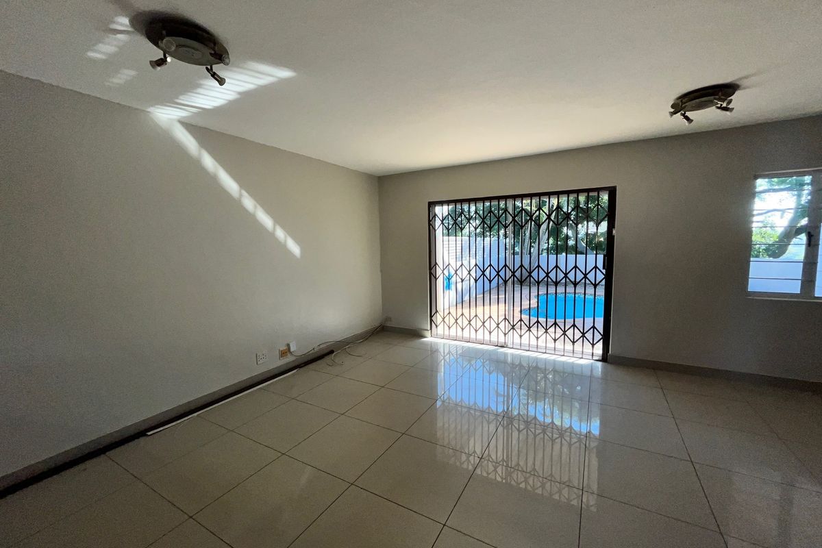 A bright, empty living room featuring large format floor tiles, ceiling lighting, and a sliding glass door with a security gate leading to the pool area.