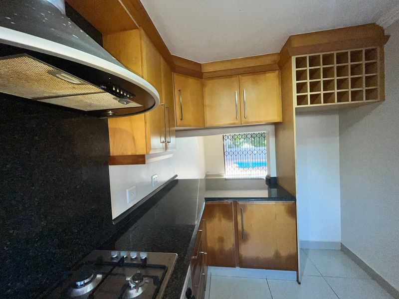 A functional kitchen featuring wooden cabinetry, granite countertops, a gas hob with extractor fan, and a built-in wine rack, with tiled flooring throughout.