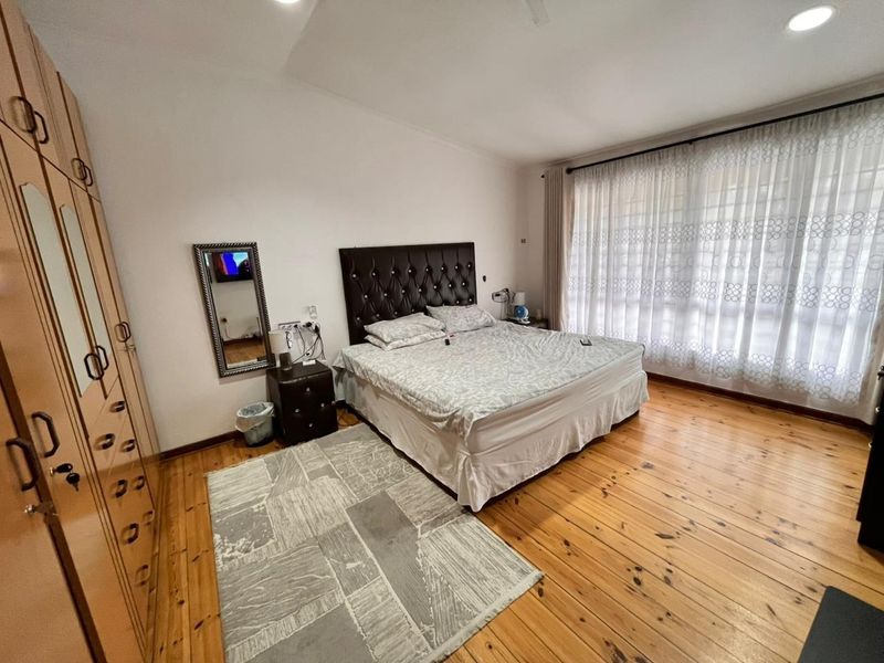 A spacious bedroom featuring warm timber flooring, built-in wooden cupboards, and large windows with sheer curtains allowing for ample natural light.