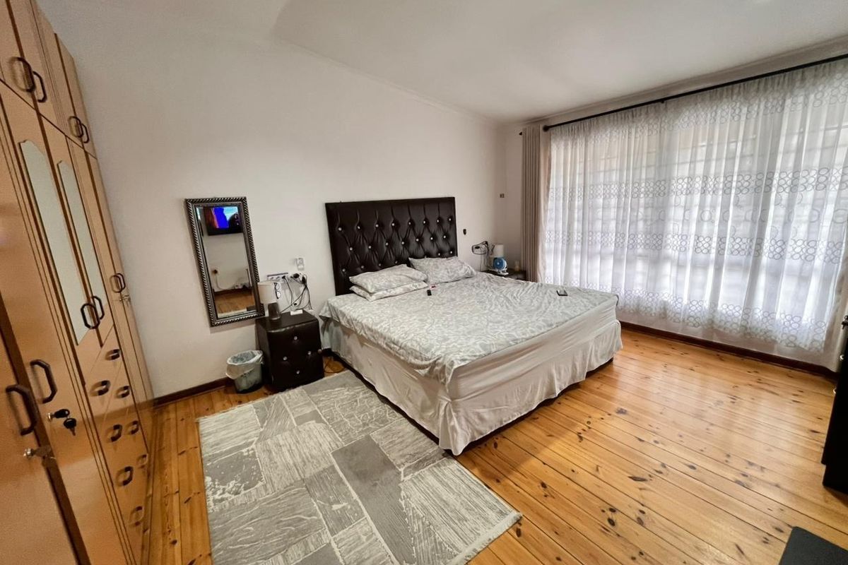 A spacious bedroom featuring warm timber flooring, built-in wooden cupboards, and large windows with sheer curtains allowing for ample natural light.