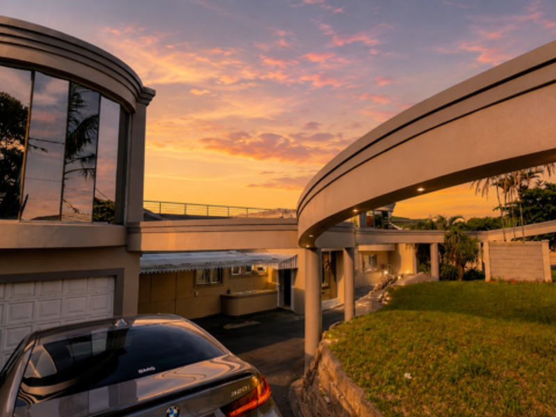 A striking modern architectural front elevation featuring a curved design, integrated lighting, and a paved driveway at sunset.