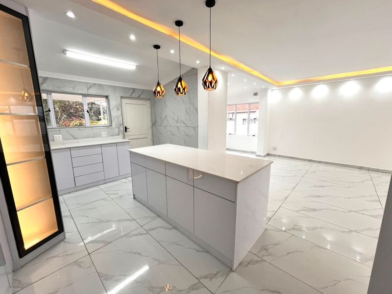 Modern open-plan kitchen and living area featuring high-gloss cabinetry, a central island with stone countertops, marble-look floor tiles, and sophisticated recessed and pendant lighting.