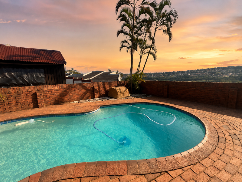 A well-maintained swimming pool area featuring brick paving and a brick boundary wall, set against a scenic sunset backdrop.