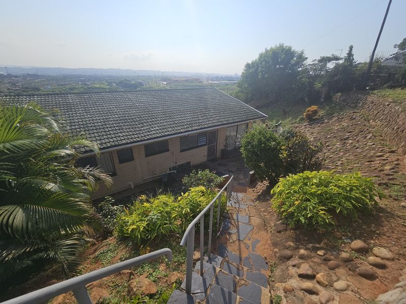Rear view of the property featuring a tiled roof, stone staircase with metal handrail, and a sloped garden area with distant landscape views.