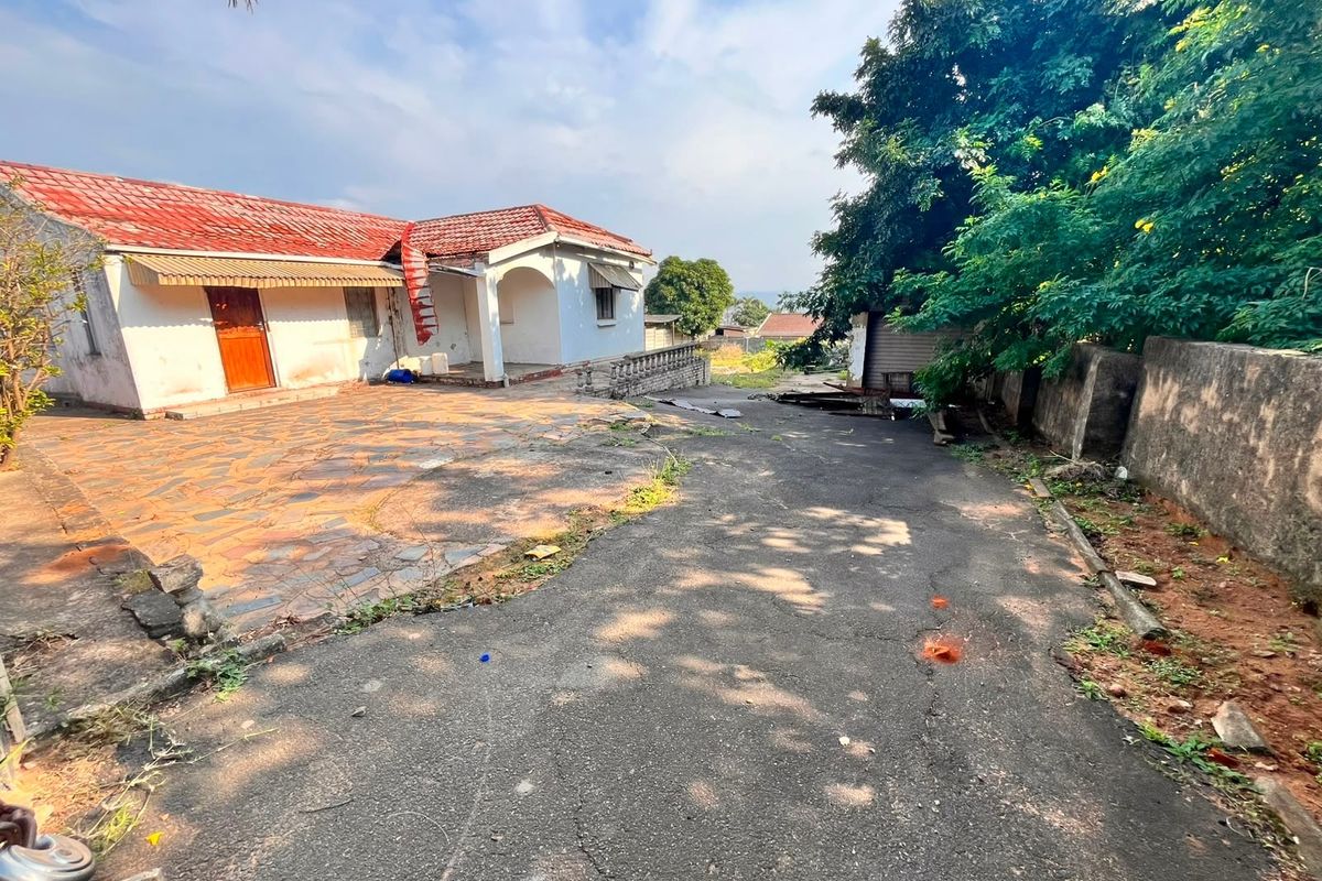 Rear exterior view of the property featuring a paved patio area, covered porch, and an asphalt driveway with surrounding garden space.