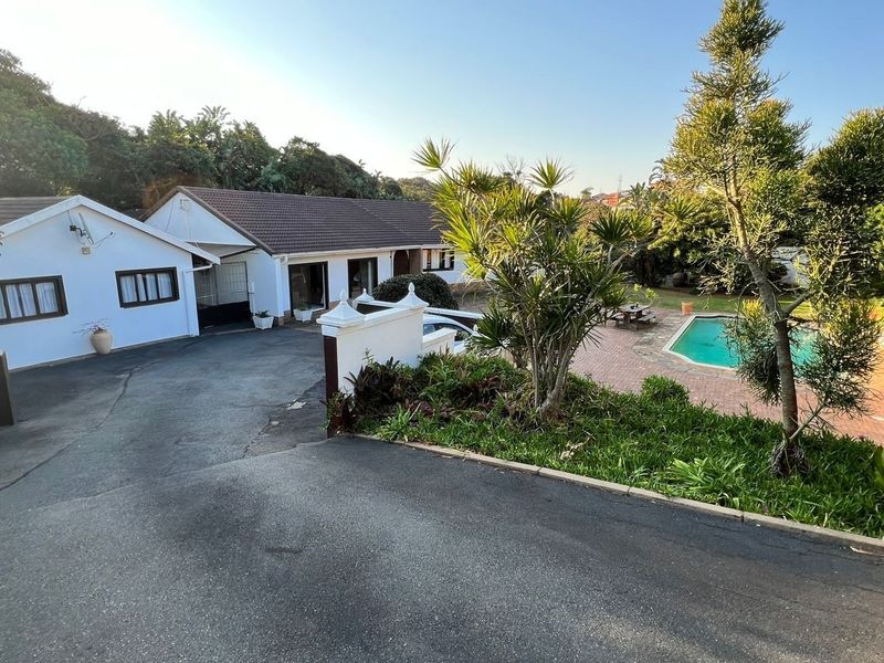 A wide-angle view of the property exterior featuring a large asphalt driveway, a swimming pool with brick paving, and established garden landscaping.