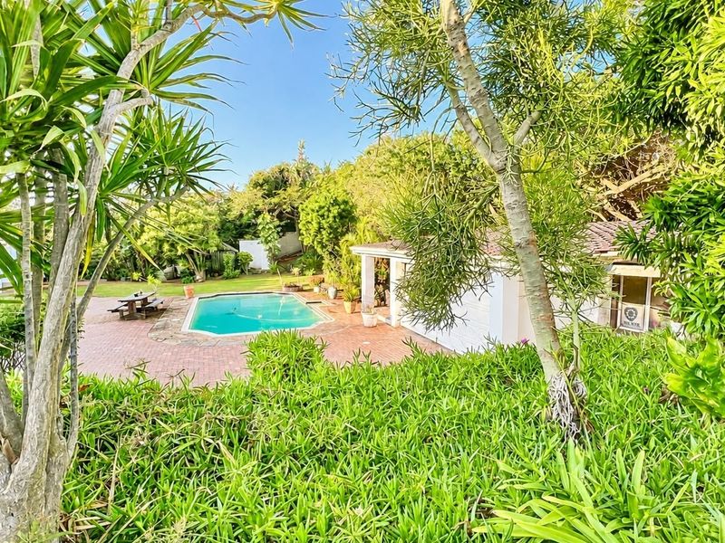 A backyard pool area featuring a swimming pool, brick paved surround, and a covered patio area set within a lush, established garden.