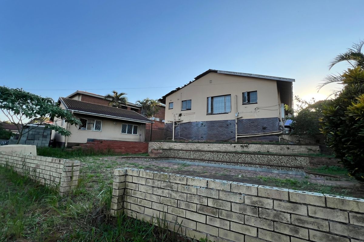 Rear view of the property featuring tiered brick retaining walls, a paved patio area, and established garden space requiring maintenance.
