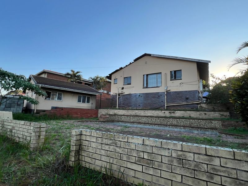 Rear view of the property featuring tiered brick retaining walls, a paved patio area, and established garden space requiring maintenance.