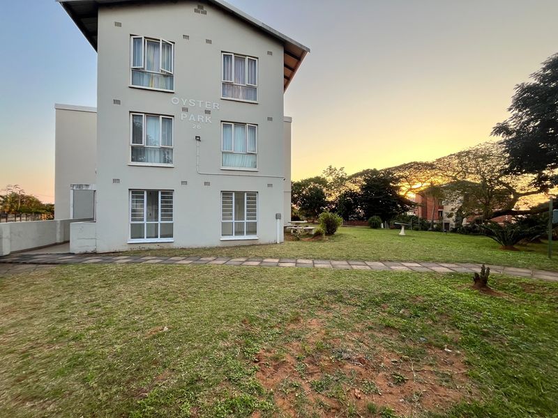 Exterior view of the Oyster Park apartment building featuring a paved walkway and surrounding communal garden area at sunset.