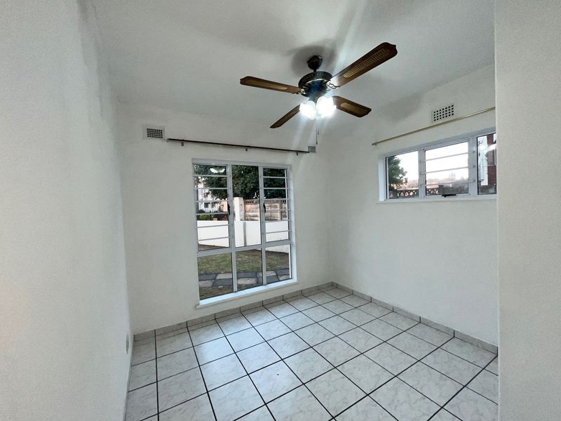 A bright, empty bedroom featuring tiled flooring, a ceiling fan, and large windows with security bars, offering a clean and neutral space.
