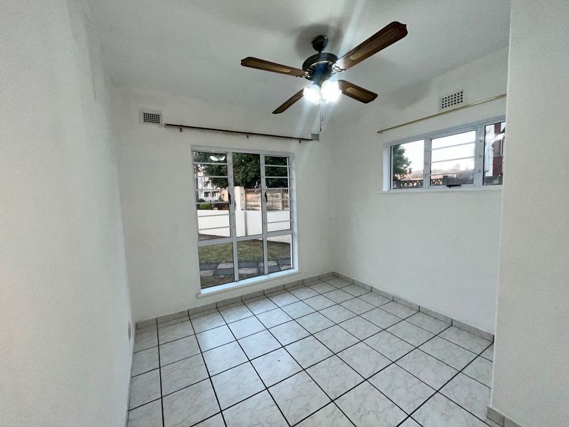 A bright, empty bedroom featuring tiled flooring, a ceiling fan, and large aluminium-framed windows providing natural light.