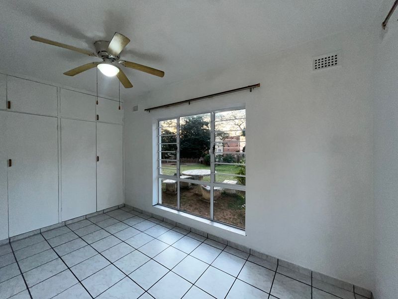 A bright, empty bedroom featuring tiled flooring, built-in cupboards, a ceiling fan, and a large window overlooking the garden.