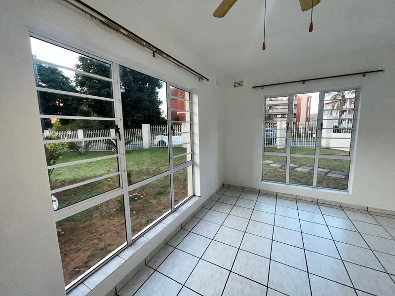 A bright, empty living area featuring tiled flooring, a ceiling fan, and large windows with security bars overlooking the garden.