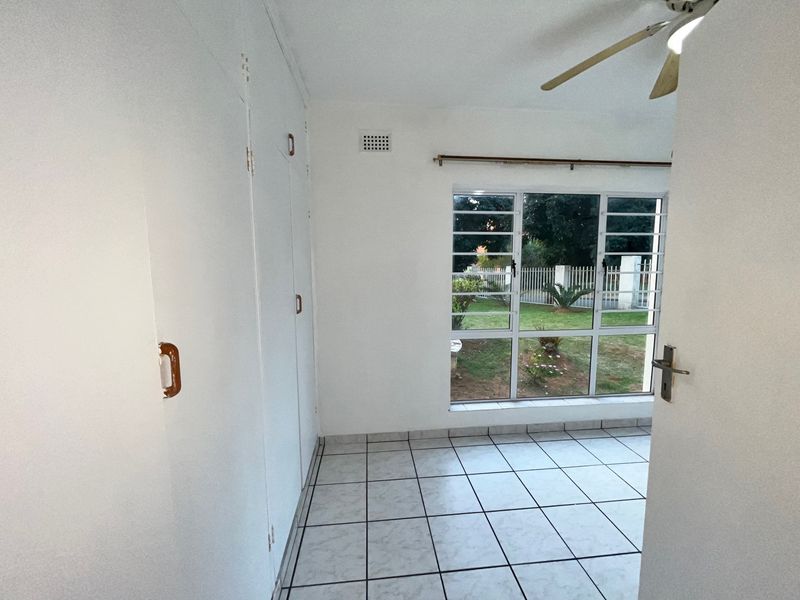 An empty bedroom featuring tiled flooring, built-in cupboards, a ceiling fan, and a large window overlooking the garden.