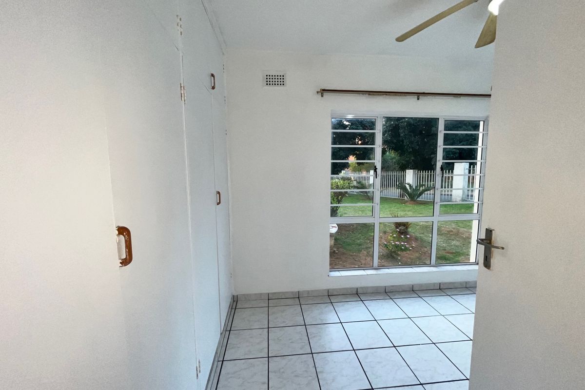 An empty bedroom featuring tiled flooring, built-in cupboards, a ceiling fan, and a large window overlooking the garden.