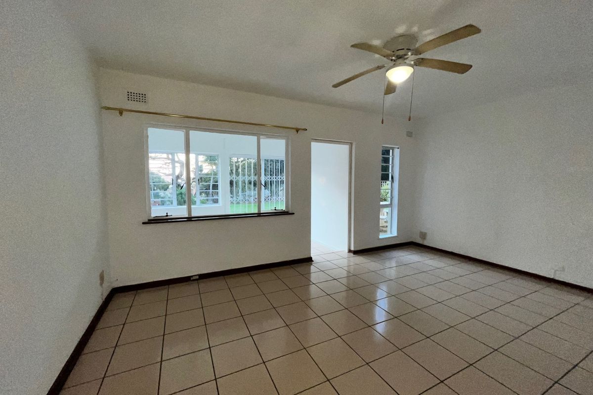 A spacious, empty living area featuring neutral tiled flooring, a ceiling fan, and large windows with security bars.