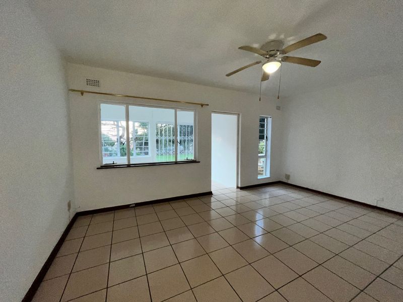 A spacious, empty living area featuring neutral tiled flooring, a ceiling fan, and large windows with security bars.