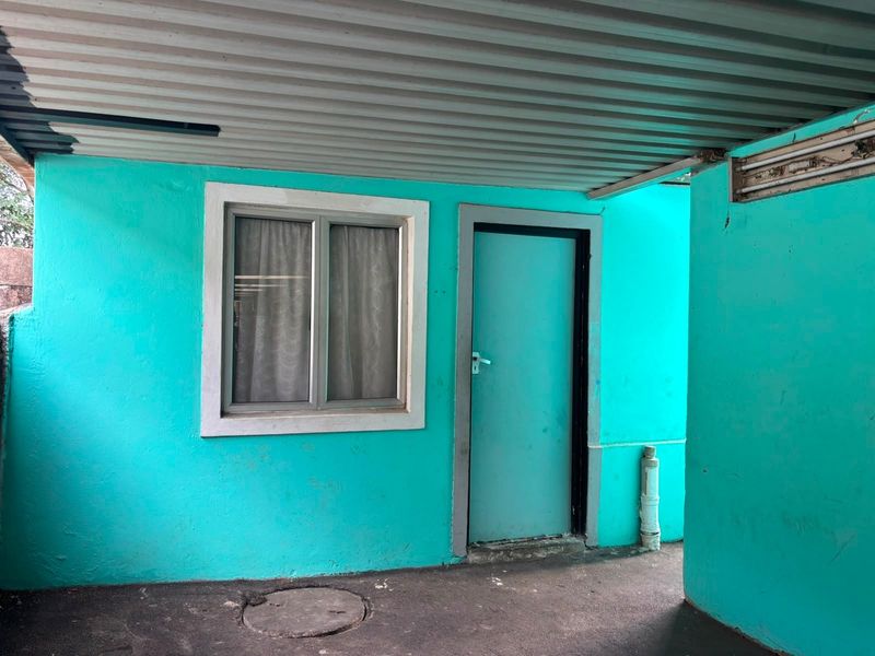 A covered rear exterior area featuring a bright turquoise painted wall, aluminium window, and solid door with concrete flooring.