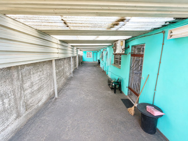 A covered outdoor side passage featuring a corrugated roof, concrete boundary wall, and direct access to the main building via a security gate.