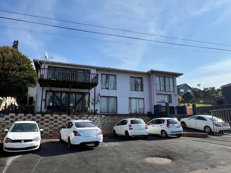 Front exterior view of a multi-story residential building featuring a paved parking area, retaining wall, and upper-level balcony with sliding door access.