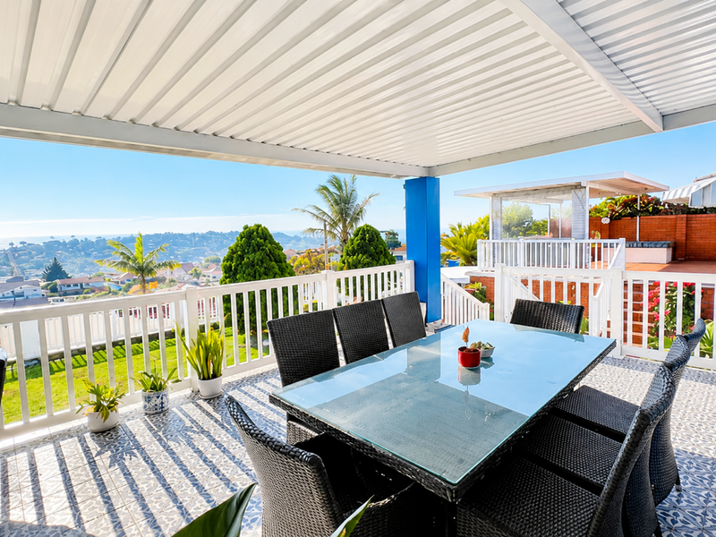 A spacious covered patio featuring patterned floor tiles, a white balustrade, and expansive elevated views of the surrounding neighbourhood and coastline.