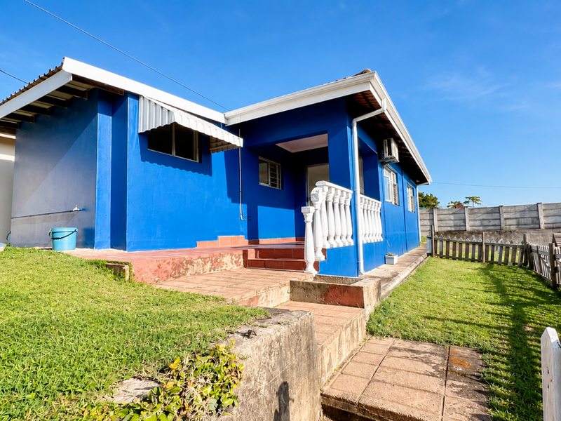 Front view of a blue-painted house featuring a tiled patio, decorative balustrade, and a surrounding garden with a timber fence and retaining wall.