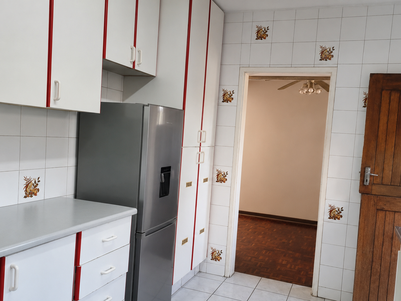 A functional kitchen featuring white cabinetry with red accents, tiled walls, laminate countertops, and a stainless steel sink, leading into an adjacent living area.