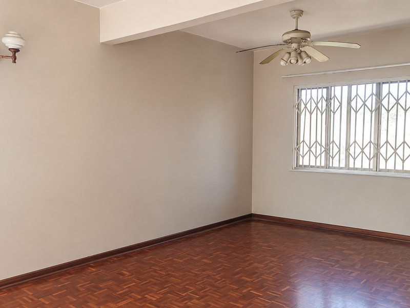 A spacious, empty living room featuring polished parquet flooring, a ceiling fan, and a window fitted with a security gate.