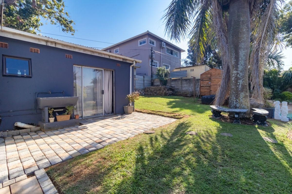 Rear garden featuring a paved patio area, built-in braai, and established lawn with mature trees and boundary walling.