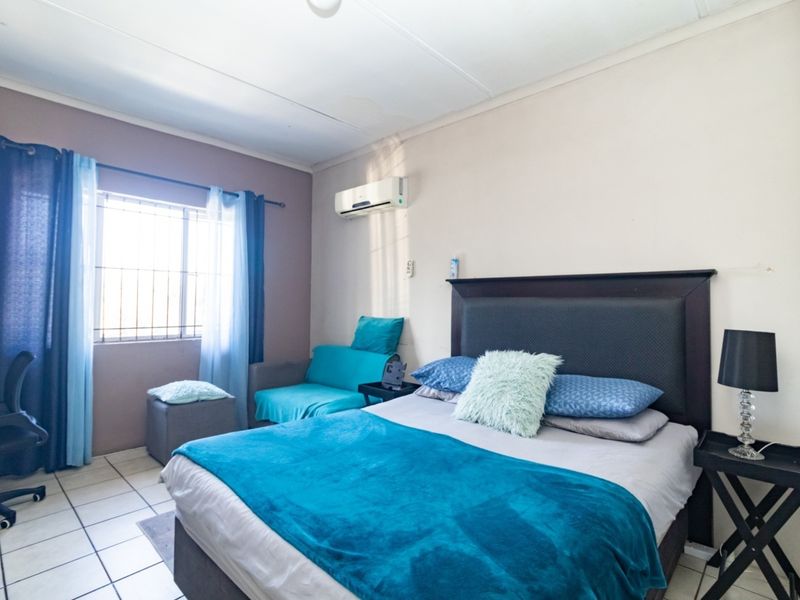 A functional bedroom featuring tiled flooring, a wall-mounted air conditioning unit, and a window fitted with security bars.