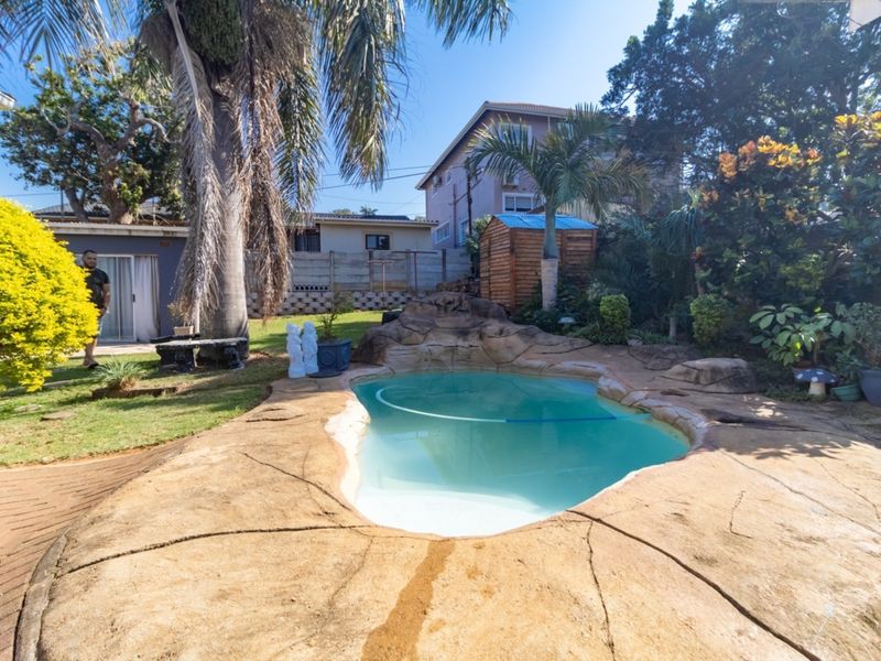 A landscaped backyard featuring a rock-style swimming pool, paved surround, and established garden area with a storage shed.