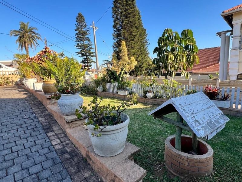 A landscaped front garden area featuring a paved driveway, a low retaining wall, a picket fence, and a decorative wishing well feature.