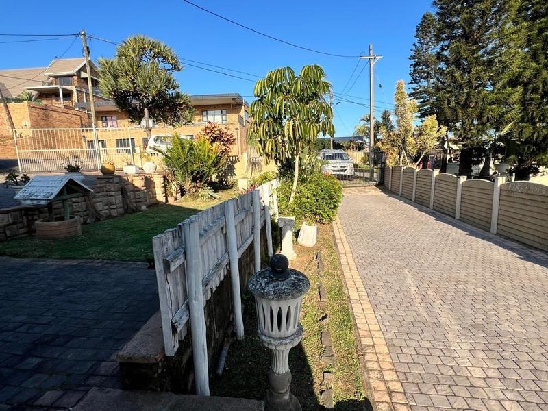 Front exterior view featuring a paved driveway, secure gated entrance, and established garden area with stone retaining walls and boundary fencing.