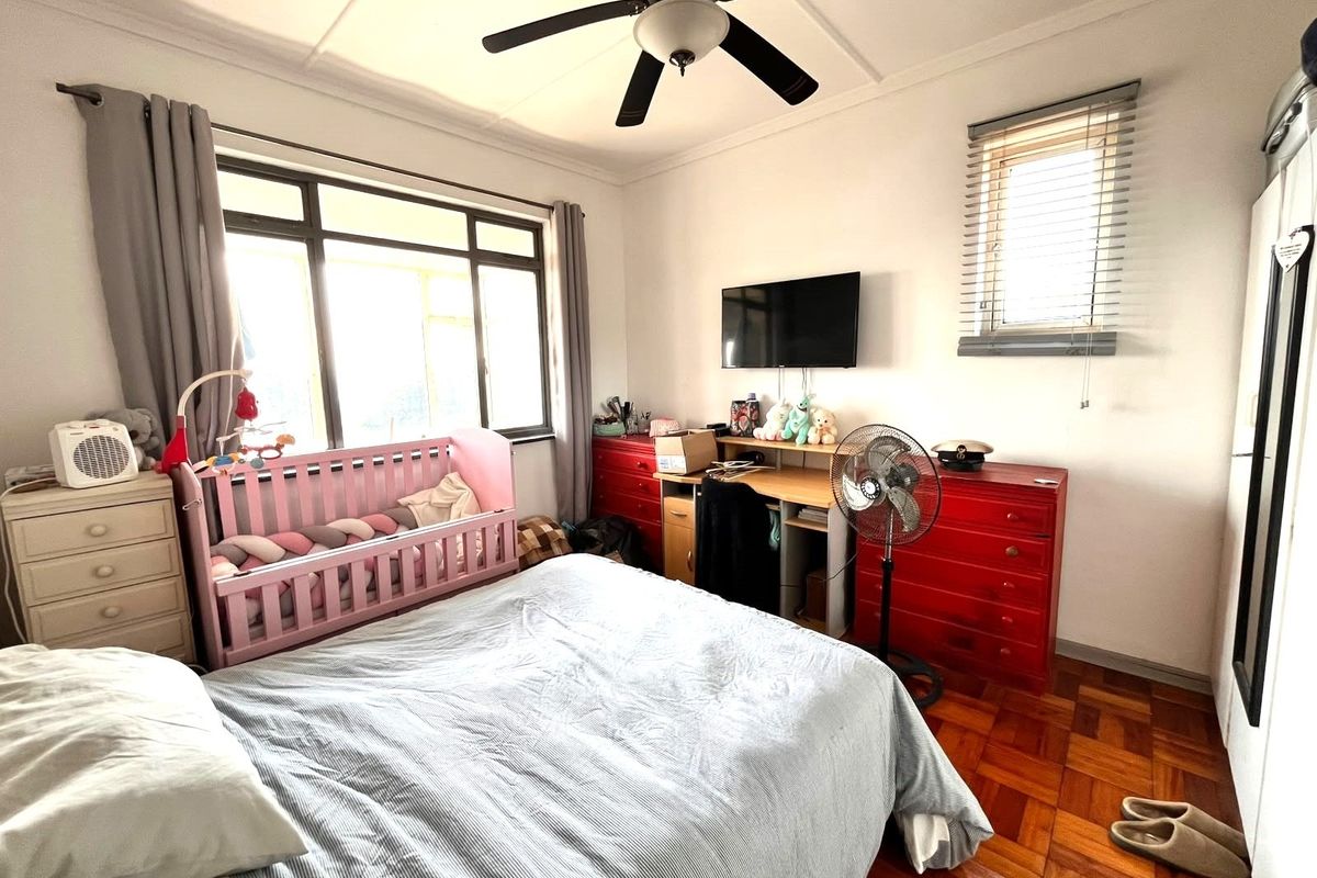 A bedroom featuring classic parquet flooring, a ceiling fan, and large windows with venetian blinds, offering a functional and well-lit living space.
